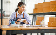 © David - Young Asian entrepreneur SME woman checking product on stock take note with a laptop. Small business owner working at home. Check address on the parcel SME online start up freelance concept.