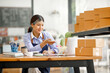 © David - Young Asian entrepreneur SME woman checking product on stock take note with a laptop. Small business owner working at home. Check address on the parcel SME online start up freelance concept.
