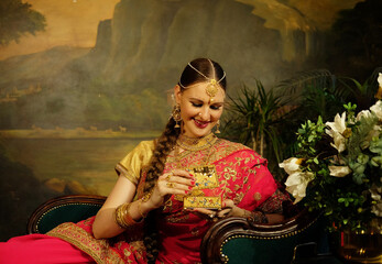 Indian bride dressed in Hindu red traditional wedding clothes sitting on sofa holding small gold box.