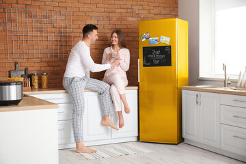 Wall Mural - Happy young couple in pajamas drinking coffee near refrigerator in kitchen