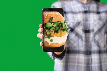 Wall Mural - Young woman with mobile phone ordering burger online against green background, closeup