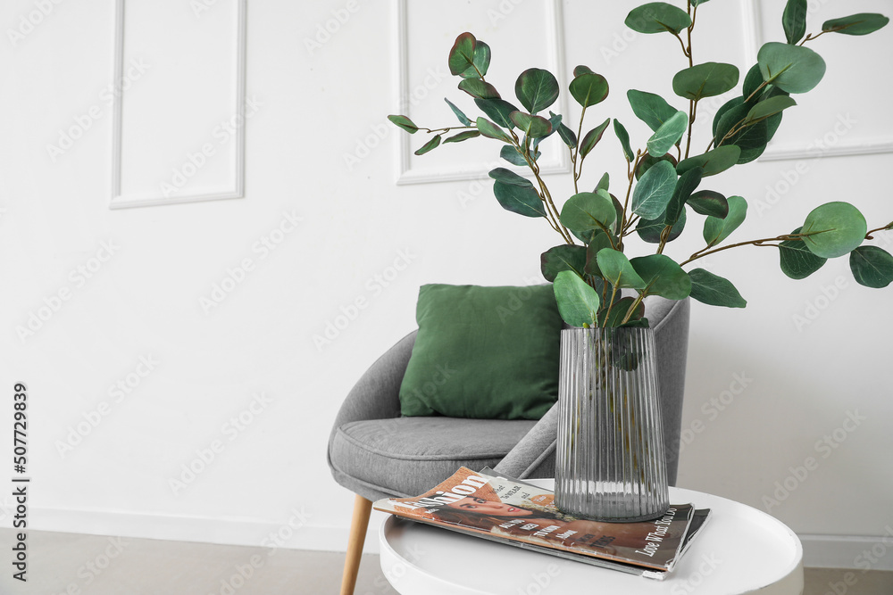 Table with eucalyptus in vase, magazine and armchair near light wall