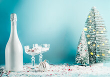 Christmas And New Year Background With White Champagne Bottle, Glasses And Fir Trees At Blue Backdrop With Snow And Bokeh. Seasonal Winter Setting With Festive Drink. Front View With Copy Space.