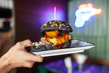 Hands Of Woman Holding Black Burger On Plate At Restaurant