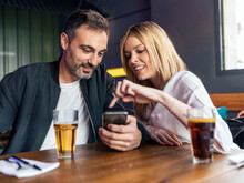 Smiling Woman Pointing At Smart Phone Held By Man Sitting At Table In Restaurant