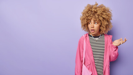 Canvas Print - Horizontal shot of confused hesitant curly woman shrugs shoulders feels uncertain wears casual striped jumper and pink jacket feels bewildered isolated over purple background blank copy space