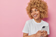 © Wayhome Studio - Smiling beautiful woman with curly bushy hair uses mobile phone chats online in messanger dressed in casual white t shirt focused away isolated over pink background blank space for your promotion