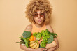 © wayhome.studio  - Surprised curly haired young woman keeps lips folded poses with green grocery fullof vitamins eats seasonal vegetables and fruits wears sunglasses and t shirt isolated over beige background.