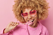 © wayhome.studio - Photo of lovely curly haired young woman bites lollipop winks eye wears trendy sunglasses has sweet tooth dressed in fashionable jacket isolated over pink background. Female model with lollipop