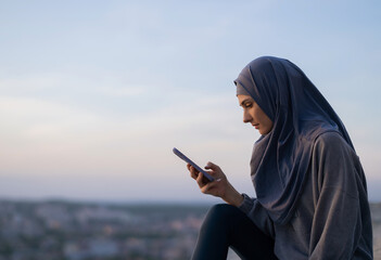 Wall Mural - Young beautiful girl in a hijab says looking at the phone on a background of sunset and cityscape