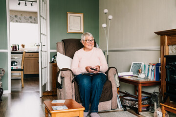 Portrait of smiling retired elderly woman in comfortable armchair with TV remote control and watching television channel in living room. Retirement concept. Selective focus.