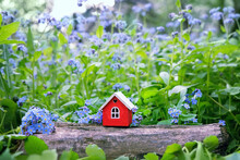 Wooden Red Toy House And Blue Flowers Of Forget Me Not In Garden, Blurred Green Natural Background. Eco Friendly Home. Symbol Of Family. Mortgage, Real Estate Concept. Spring Summer Season