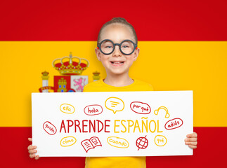 Beautiful cute little girl holding a white board with Learn Spanish text in Spanish and illustrations in front of blurry flag of Spain. English: Learn Spanish. Foreign language learning concept.
