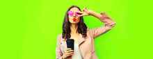 Portrait Of Beautiful Young Woman Blowing Her Lips Sending Air Kiss Posing Wearing Pink Sunglasses, Leather Jacket On Green Background