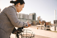Side View Of Businesswoman With In-ear Headphones Using Smart Phone While Leaning On Bicycle