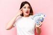 © Asier - Young caucasian woman holding banknotes isolated on pink background showing a disappointment gesture with forefinger.
