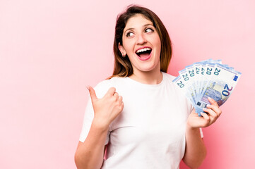 Young caucasian woman holding banknotes isolated on pink background points with thumb finger away, laughing and carefree.