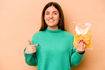 Wall Mural - Young hispanic woman holding a bag of chips isolated on beige background person pointing by hand to a shirt copy space, proud and confident