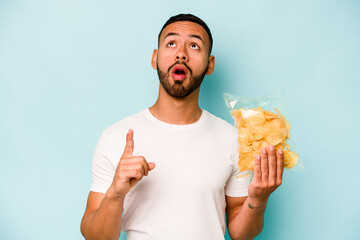 Wall Mural - Young hispanic man holding a bag of chips isolated on blue background pointing upside with opened mouth.