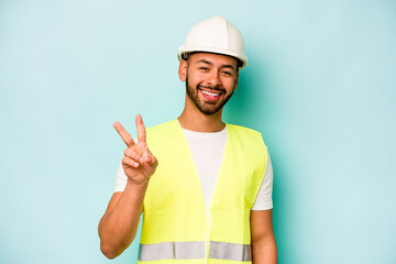 Wall Mural - Young laborer hispanic man isolated on blue background joyful and carefree showing a peace symbol with fingers.