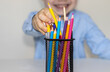 joyful kid takes a pencil from metal net box pen holder in front of him. back to school concept.cute little boy in blue shirt.elementary school or homeschooling, distance learning. colorful pencils