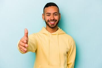 Wall Mural - Young hispanic man isolated on blue background stretching hand at camera in greeting gesture.