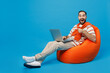 Full body young smiling cheerful fun man 20s wear orange striped t-shirt sit in bag chair hold use work point index inger on on laptop pc computer isolated on plain blue background studio portrait