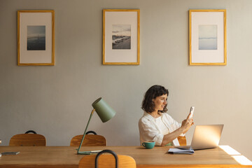 Beautiful young caucasian woman uses modern smartphone sitting at her desk in office. Brunette in casual clothes is browsing social media. Concept of positive emotions 