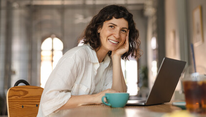 Happy young caucasian girl looks into camera sitting at table with laptop and cup of tea in cafe. Brunette wearing white shirt is working on project. Programmer concepts 