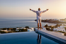 A Happy Man With A Drink Stands By The Swimming Pool And Enjoys The View Over The Sea During A Summer Sunset