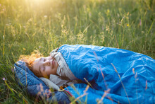 A Child Sleeps In A Sleeping Bag On The Grass In A Camping Trip - Eco-friendly Outdoor Recreation, Healthy Lifestyle, Summer Time. Sweet And Peaceful Sleep. Mosquito Bites, Repellent.
