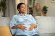 © Prostock-studio - Joyful Pregnant Black Lady Touching Belly Sitting On Sofa Indoors