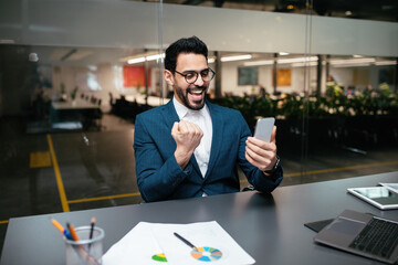 Wall Mural - Glad emotional young middle eastern businessman with beard in glasses and suit looks at smartphone