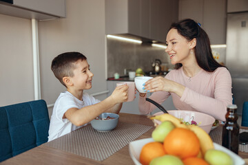 Pretty Caucasian mother and little son doing cheers gesture with cups of tea and coffee.