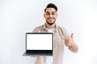 © Kateryna - Joyful arabian or indian guy in casual wear, freelancer or student, holding an open laptop with empty mock-up space and points finger on it, stand on white isolated background, looks at camera, smiles