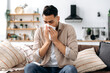 © Kateryna - Unhappy sad indian or arabian guy suffering from fever and flu, sitting on sofa in living room, blowing nose and sneezing in napkin, need treatment of illness, and doctors consultation