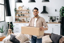 Joyful Stylish Indian Or Arabian Guy, Stand At Home In The Living Room, Holding A Large Cardboard Box, Received A Long-awaited Parcel From The Online Store, Preparing To Unpack, Looks At Camera,smiles