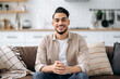 © Kateryna - Portrait of an attractive handsome indian or arabian guy in a stylish casual wear, sitting on sofa at home in a living room, in a stylish interior, looking at the camera, smiling positively