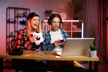 Wall Mural - Overjoyed blond woman in cap and her young man in headset gesturing and screaming while looking at screen of laptop. Young caucasian couple celebrating win of favorite team.