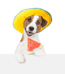 Happy jack russell terrier puppy wearing summer hat holds a watermelon in it paw above empty white banner.  isolated on white background