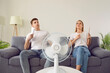 © Studio Romantic - Young couple at home suffering from hot sultry sweltering summertime weather. Sweaty exhausted people waving sheets of paper sitting on sofa with electric fan in overheated house. Summer heat concept