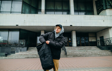 A young man in warm casual clothes stands on the street in the background of the building and pulls things out of his backpack and looks away.