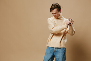 a handsome attractive guy in a long beige sweater stands straightening his sleeves with his face turned away from the camera on a monophonic studio background with empty space