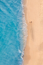 Aerial View Of Couple Walking On Beach With Sunlight Close To Turquoise Sea Waves. Top View Of Summer Beach Landscape, Romantic Inspirational Couple Vacation, Romance Holiday. Freedom Travel Template