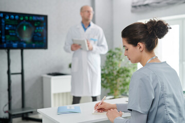 Young student in uniform sitting at desk and making notes while doctor holding lecture at class