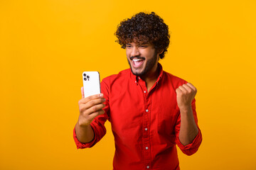 Happy satisfied man with beard holding smartphone and smiling making yes gesture, celebrating online lottery or giveaway victory. Indoor studio shot isolated on orange background
