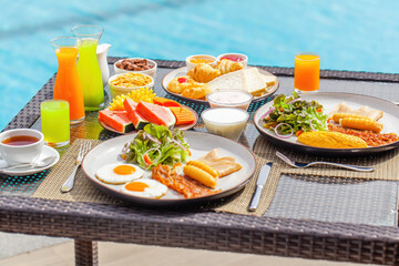 Wall Mural - Hotel breakfast. Pair having breakfast on tropical villa near swimming pool. Table full of food - exotic fruits, eggs and bakery at luxury hotel in hot summer day. Balanced food