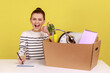 Excited woman office worker sitting at workplace with cardboard box with her things and smiling, showing thumb up, signs an agreement. Indoor studio studio shot isolated on yellow background.