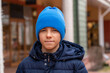 © sommersby - Portrait of confident teen boy in warm coat and blue hat looks directly into camera at city street.