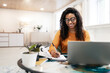 © Prostock-studio - Woman sitting at desk, using computer and writing in notebook